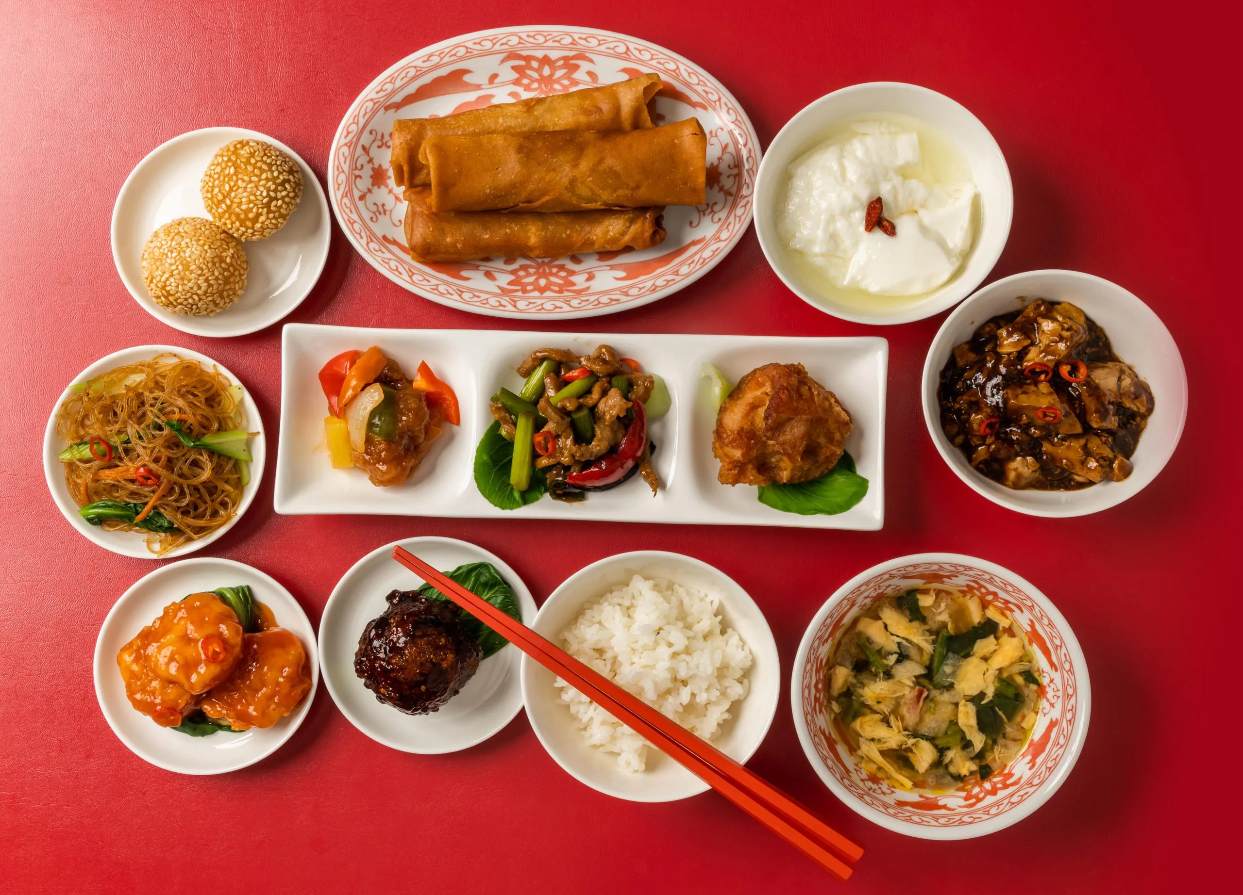 A festive spread of Chinese dishes on a red background, including spring rolls, sesame balls, rice, soup, stir-fries, and assorted proteins in white and patterned porcelain.